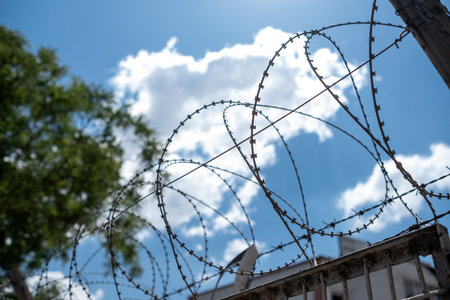 Barbed wire coils twist against a vibrant blue sky adorned with clouds, suggesting security measures...
