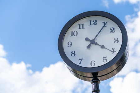 A clock mounted on a pole displays the time under a bright blue sky with fluffy white clouds,...