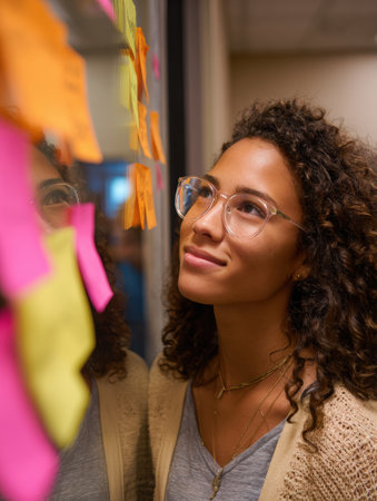 A student observes her reflection in a mirror, thoughtfully looking at sticky notes displaying her goals, in a bright academic space.の素材