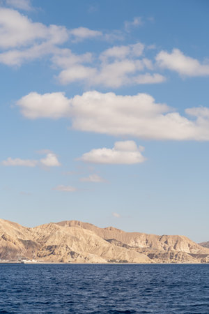 Mountains rise majestically above calm waters, with a few clouds floating in a bright blue sky. This...
