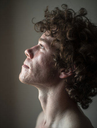 A young man with curly hair gazes upward, the soft light highlighting his facial features, creating a contemplative and serene atmosphere in a muted background.の素材