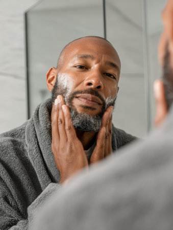 A man carefully applies soap to his beard while looking in the mirror, dressed in a soft robe in a bright bathroom during morning hours.の素材