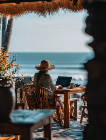 Worker sits at a wooden table on a deck overlooking the ocean, enjoying the serene environment and sunny weather while using a laptop.の素材