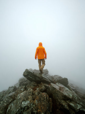 A person wearing an orange jacket walks carefully across large rocks in a misty mountain environment, illustrating a challenging hiking adventure.の素材