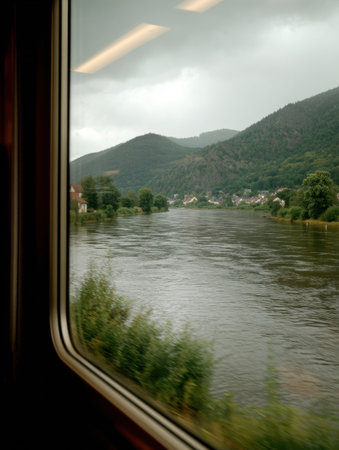 A traveler enjoys a picturesque view of a river framed by lush mountains and quaint houses while riding on a train during overcast weather.の素材