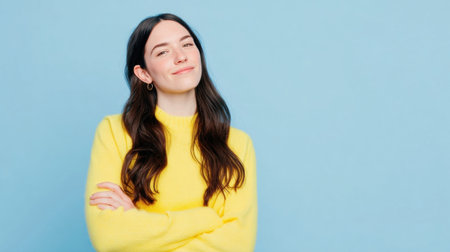 A woman with long hair and a cheerful smile stands with her arms crossed, dressed in a bright yellow sweater, set against a solid light blue background.の素材