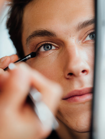 Close-up view of a young man focused on applying eyeliner in a well-lit studio, showing intricate makeup techniques and personal grooming.の素材
