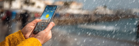 A person uses a smartphone to check the weather by the seaside while rain falls, contrasting the sunny forecast displayed on the screen.の素材