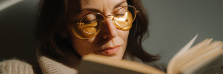 A woman enjoys her time reading a book while wearing golden under-eye patches, illuminated by warm, soft lighting in a cozy environment.の素材