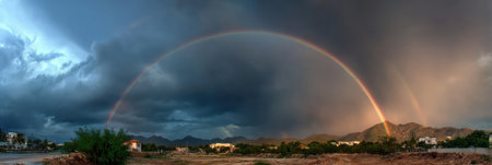 A vibrant rainbow spans the sky above a desert landscape, illuminated by evening light and contrasting with dark storm clouds, revealing natures wonders.の素材