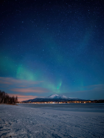 Vibrant auroras dance across the night sky above a snowy lake, with a mountain backdrop and distant lights from a nearby town creating a magical atmosphere.の素材