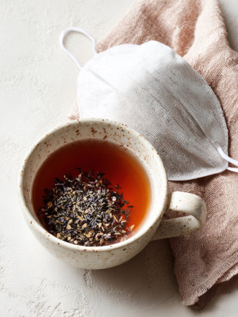 A speckled cup holds warm herbal tea with visible herbs floating atop. A soft cloth mask rests nearby, creating a serene, restful atmosphere.の素材