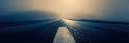 A desolate road disappears into a thick fog at dawn, with a reflective surface accentuating the tranquil yet eerie ambiance of the early morning.の素材