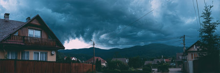 An ominous sky filled with dark storm clouds looms over a peaceful village, offering a dramatic contrast to the surrounding mountains at dusk.の素材
