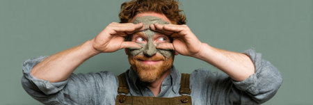 An artist displays playful creativity by shaping clay into a mask while smiling and posing with hands in a workshop filled with natural light.の素材