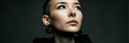 A young woman with a shaved head and stylish jewelry gazes upward, expressing confidence and creativity in a minimalist setting with dark tones.の素材