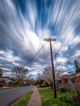 A serene suburban street showcases blooming trees alongside homes, with dynamic clouds moving across the sky, creating a tranquil yet lively atmosphere.の素材