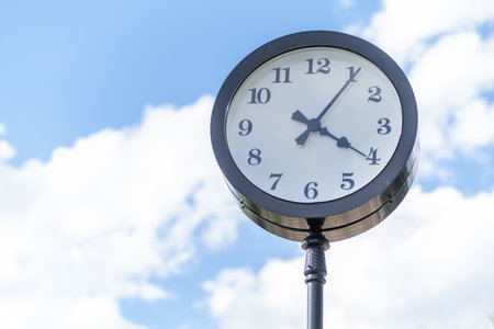 Clock mounted on a pole displays the hour clearly against a bright blue sky dotted with white...