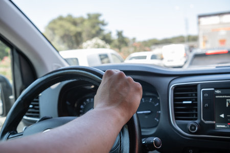 Hand gripping the steering wheel while navigating through busy city traffic on a clear, sunny day,...