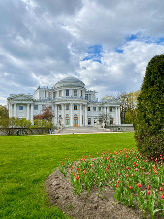 A grand mansion with classical architecture stands elegantly amid a grassy area, accented by blooming tulips and under a partly cloudy sky.の写真素材