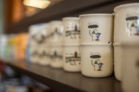 Mugs with playful illustrations are neatly arranged on a wooden shelf in a warm, inviting cafe during a relaxed afternoon.の写真素材