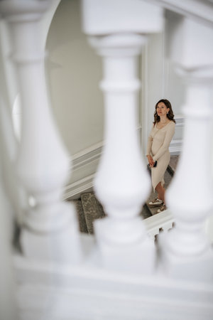 A woman wearing a fitted beige dress stands on a staircase, framed by ornate white balusters, in a bright and stylish interior.の写真素材