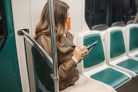 A woman dressed in a brown jacket and sitting alone on a subway train is focused on her smartphone, absorbed in her digital world during transit.の写真素材