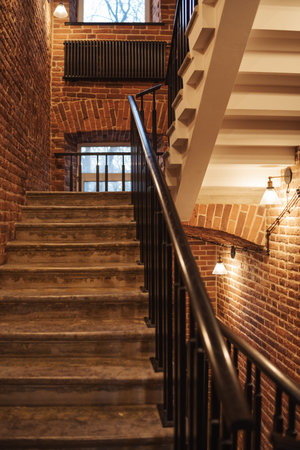 A staircase with wooden steps winds upward inside a historic building, accentuated by warm brick walls and tasteful lighting, creating a cozy atmosphere.の写真素材