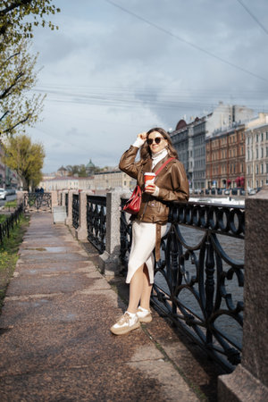 A casually dressed woman stands by a riverfront, sipping coffee while enjoying the pleasant weather of a sunny afternoon in an urban setting.の写真素材