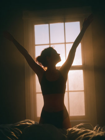 A woman stretches her arms wide in the morning light pouring through a window while standing on a bed in a serene bedroom setting.の素材