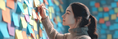 A woman writes on a sticky note while surrounded by a vibrant display of colorful notes on a wall, engaged in a productive brainstorming session.の素材