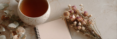 A warm cup of tea sits beside an open notebook and dried flowers on a marble tabletop, creating a serene and inviting workspace atmosphere.の素材