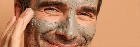 A man enjoys self-care by applying a mud mask to his face, promoting relaxation and skincare in a comfortable indoor setting during the day.の素材