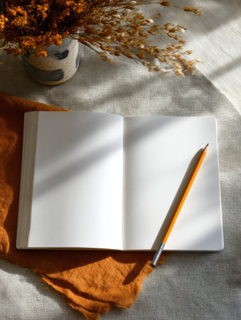 A blank notebook lies on a textured surface with a pencil beside it, while a vase of dried flowers adds a warm touch in soft natural lighting.の素材