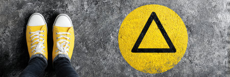Bright yellow sneakers rest on a rough surface marked by a yellow circle and black triangle, suggesting a safety or caution signal in an urban environment.の素材