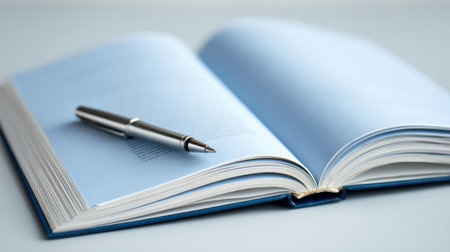 An open book lies on a flat surface, featuring blank pages while a silver pen rests beside it, offering an opportunity for writing and creativity.の素材
