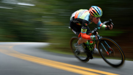Rider leans into a sharp turn on a winding road surrounded by trees, showcasing skill and speed during a competitive cycling event.の素材