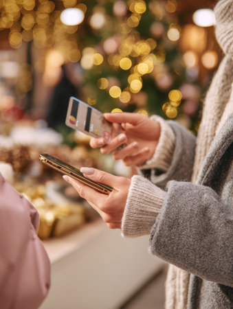 A shopper is focused on their phone while browsing at a market filled with holiday decorations and soft, glowing lights, creating a warm atmosphere.の素材