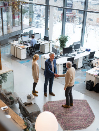 Two business professionals shake hands in a contemporary office while a third person works at a desk.の素材