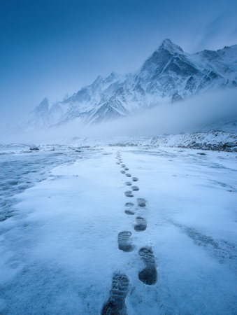 Footprints traverse a frozen landscape, guiding toward towering mountains shrouded in mist during the twilight hours, creating a serene wilderness atmosphere.の素材