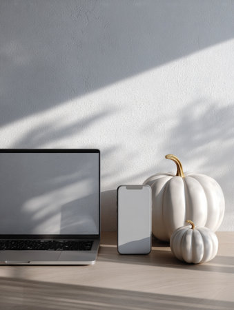 A sleek laptop and a smartphone sit on a light wooden surface alongside elegant white pumpkins under soft natural light, creating a cozy autumn atmosphere.の素材