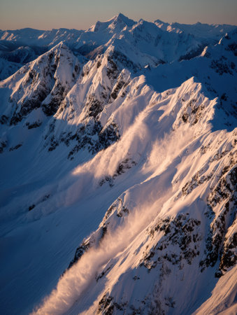 Winds sweep across a majestic mountain range, sending snow plumes into the air as the sun sets, casting golden hues on the peaks and valleys.の素材