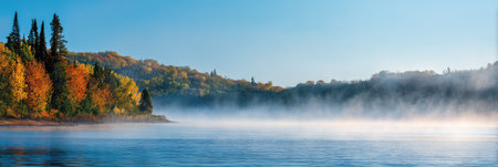 Mist rises from a calm lake as colorful autumn trees reflect the sunlight in a peaceful natural setting during early morning hours.の素材