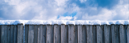 A wooden fence covered in fresh snow stands against a backdrop of bright, fluffy clouds and a clear blue sky, showcasing a serene winter day.の素材
