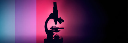 A microscope stands in silhouette against a colorful background of pink and purple lighting, highlighting its shapes and components in a laboratory ambiance.の素材