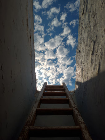 A wooden ladder leads upward from a dim space, framed by walls, revealing a bright sky filled with fluffy clouds on a sunny day.の素材