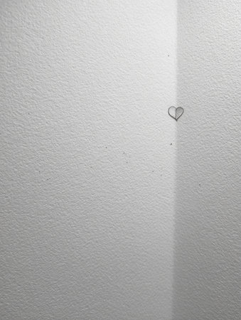A delicate heart shape formed by a paper clip is fixed to a plain white wall, adding a touch of warmth to the interior space in soft afternoon light.の素材