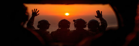 Four soldiers in military attire, wearing headsets, are standing inside a helicopter. They raise their hands and wave as the sun sets over the ocean.の素材