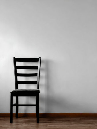 A solitary black wooden chair stands on wooden flooring beside a plain white wall, illuminated by natural light from a nearby window.の素材