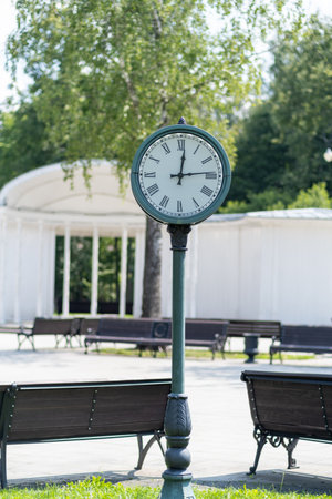 A traditional clock mounted on a pole is positioned in a park filled with green grass and empty benches under a clear blue sky during daylight.の写真素材